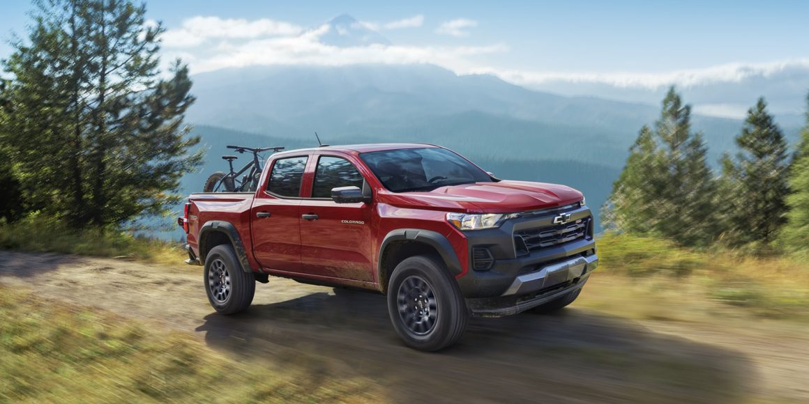 Red 2026 Chevrolet Colorado crew cab carrying a mountain bike on a dirt trail with mountains in the background