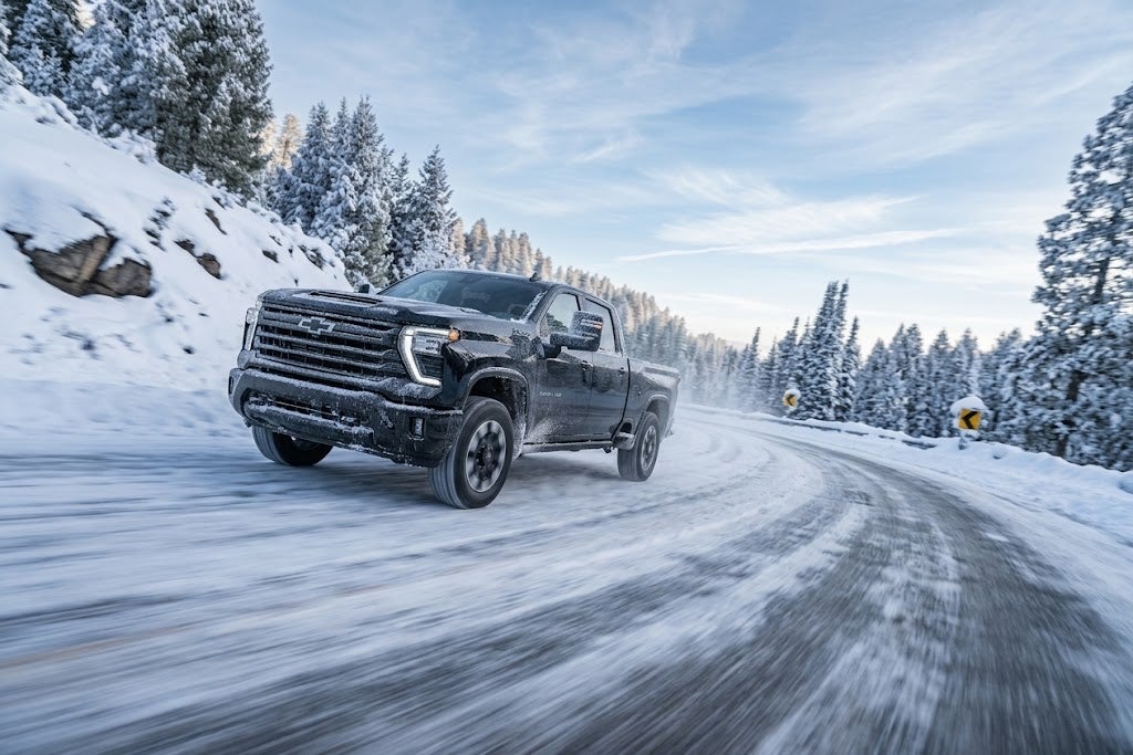 Black Chevy Silverado HD pickup truck driving on a snow-covered road lined with frosted evergreen trees in winter